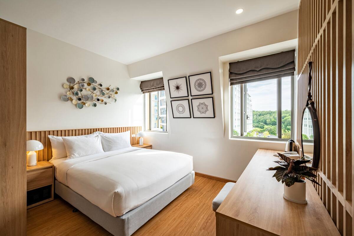 Une chambre moderne avec un grand lit, des tables de chevet avec des lampes, une tête de lit en bois, un bureau avec une plante et une vue sur la ville à travers de grandes fenêtres.