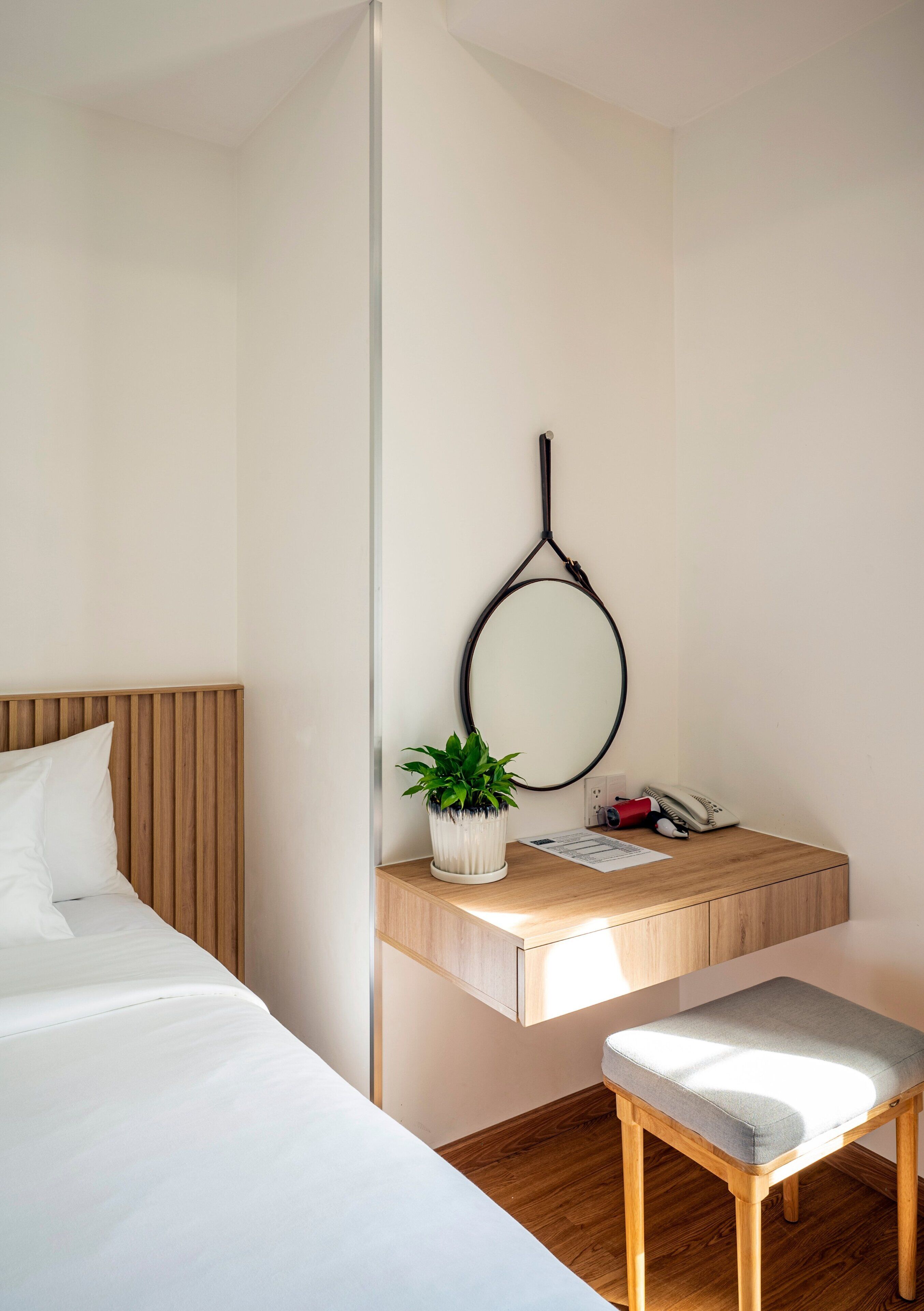 A modern hotel room with a bed, a wooden bedside table with a plant, a mirror, and a telephone.