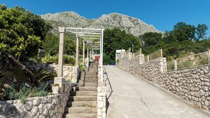 A stone staircase leading uphill with a metal railing on one side and a stone wall on the other, surrounded by greenery and a mountain in the background. - Villa Barbara (Zupa dubrovacka)