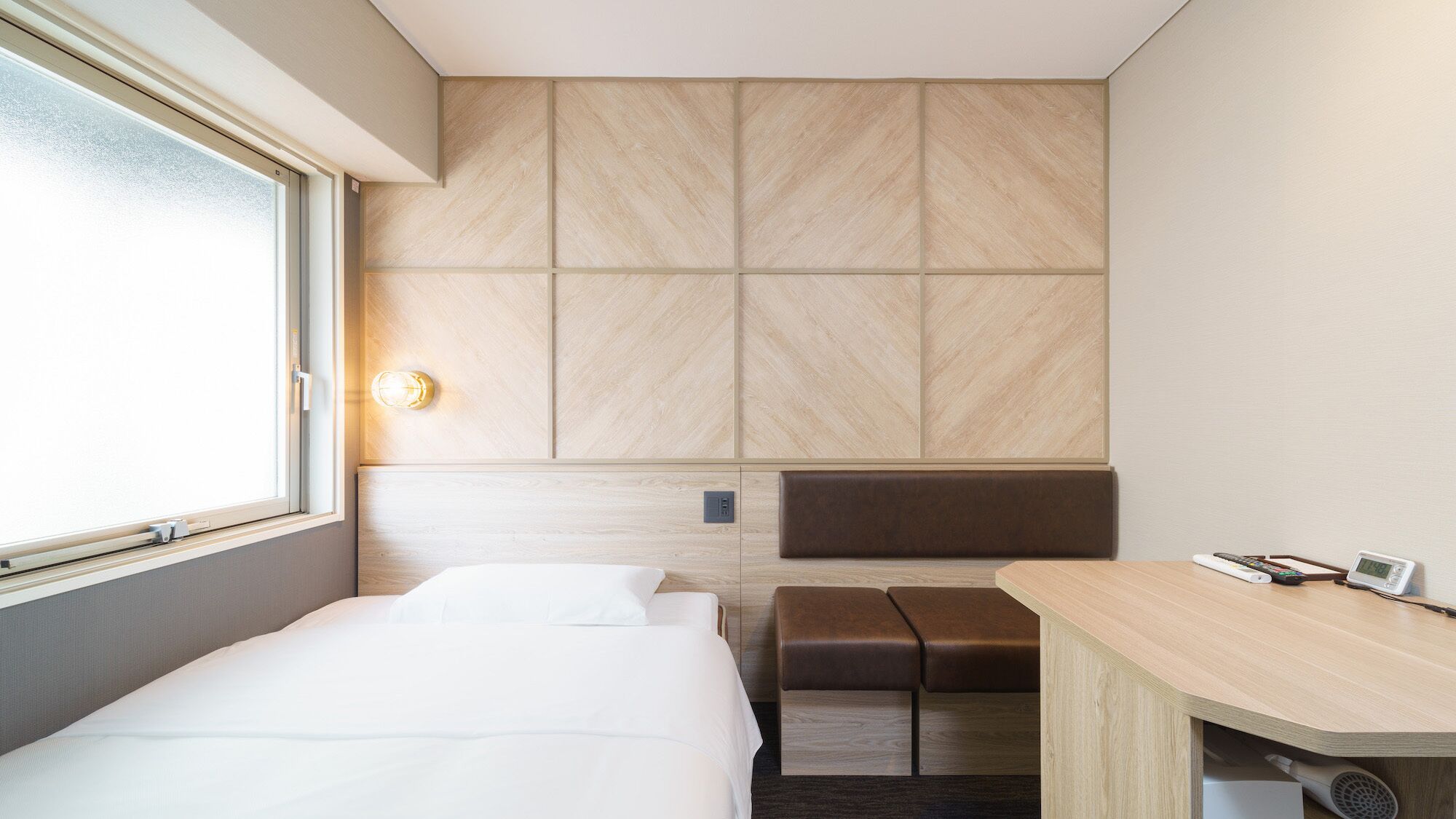 A hotel room with a bed, a desk with a chair, a bench, and a large window.