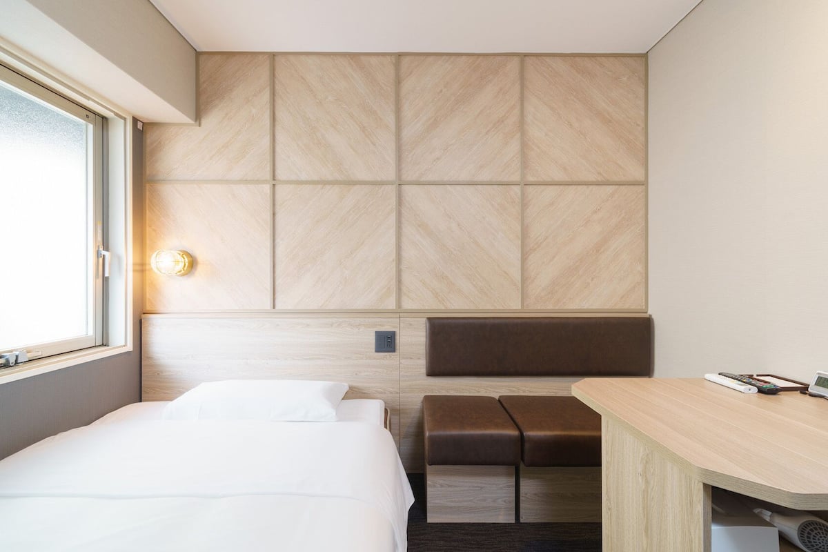 Een hotelkamer met een bed, een bureau met stoel, een bankje en een groot raam.