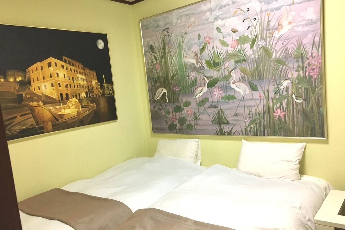 A hotel room with a bed, a night view painting of a building, and a nature scene painting.