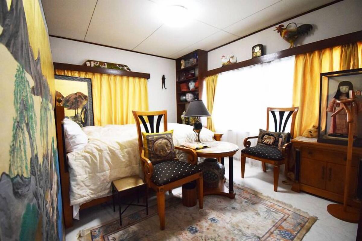 A bedroom with a bed, two wooden chairs, a small table, a lamp, a mirror, and a shelf with decorative items.