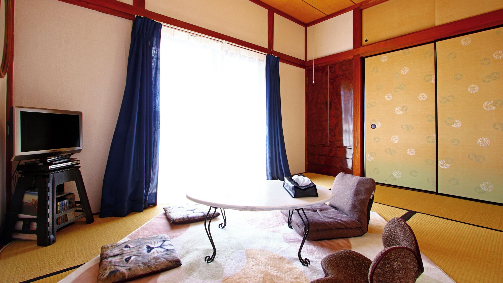 Quarto japonês tradicional com piso de tatame, uma televisão em um suporte, uma mesa e uma porta deslizante com papel estampado.