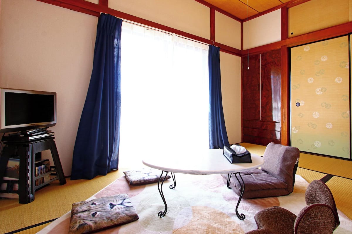 Una habitación japonesa tradicional con suelo de tatami, un televisor sobre un soporte, una mesa y una puerta corredera con papel decorado.
