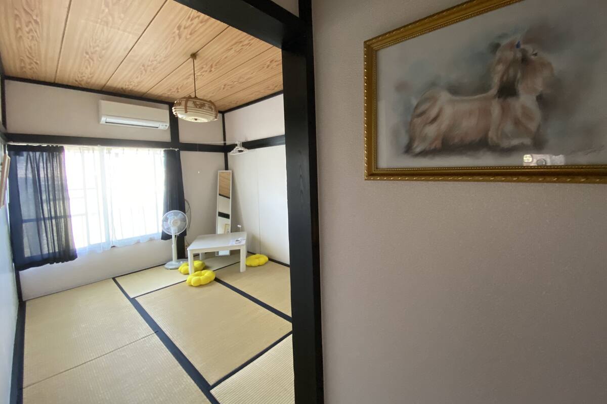 Una habitación con suelo de tatami, un escritorio blanco, un ventilador de pie y un cuadro en la pared.