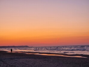 Escena de playa con una persona de pie en la arena, olas acercándose a la orilla y un atardecer al fondo.