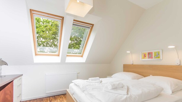 Et soveværelse med skråvæg, to vinduer med udsigt til træer, en træsenggavl, et natbord og en radiator.