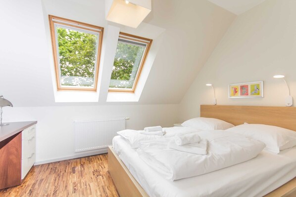 A bedroom with a sloped ceiling, two windows with a view of trees, a wooden headboard, a bedside table, and a radiator.