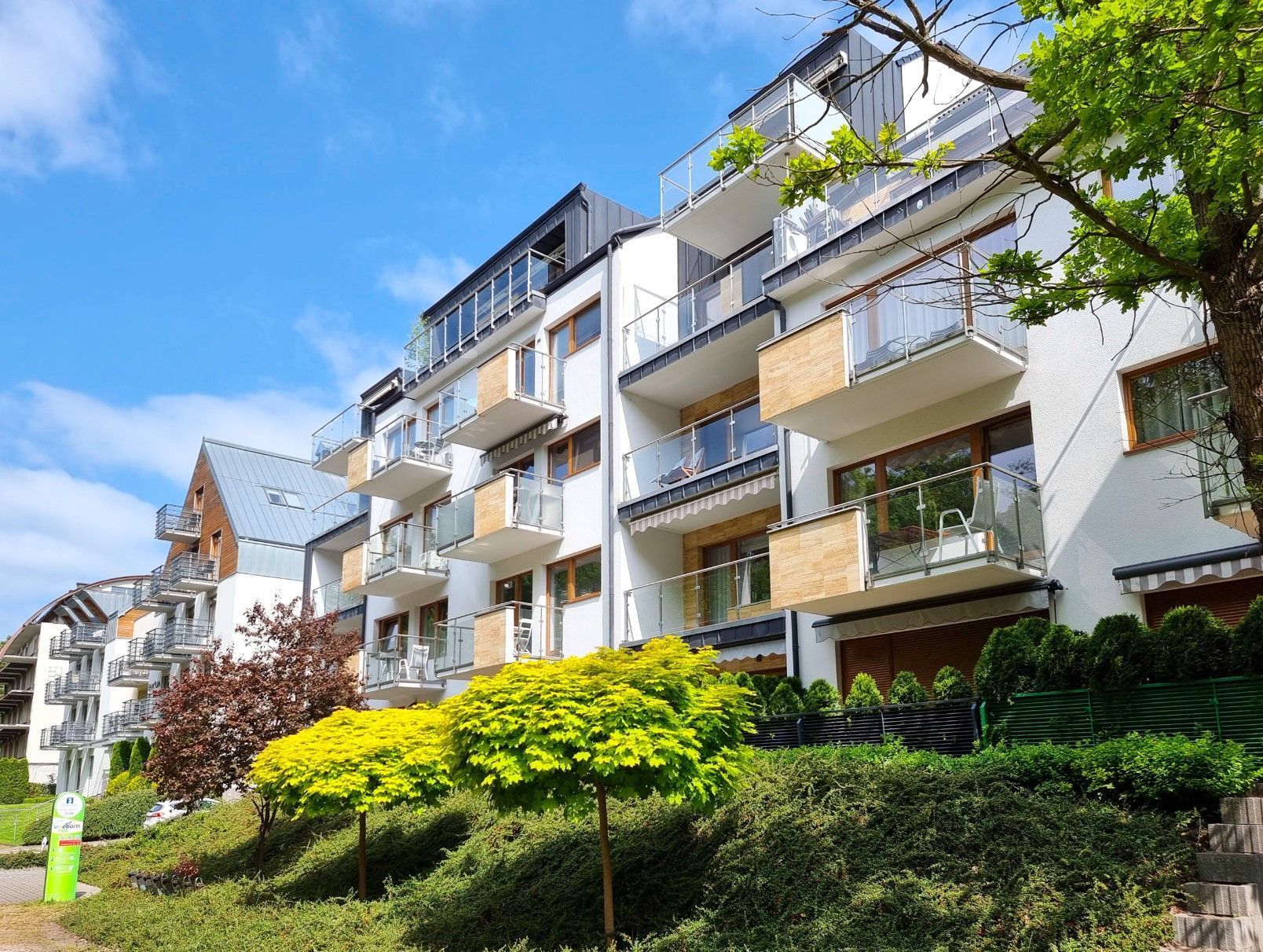 Un immeuble résidentiel à plusieurs étages, doté de balcons, entouré d’un espace vert aménagé en parc, et surplombant un ciel dégagé.