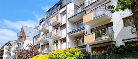 Un immeuble résidentiel à plusieurs étages, doté de balcons, entouré d’un espace vert aménagé en parc, et surplombant un ciel dégagé.