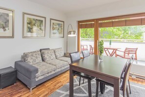 A living room with a grey sofa, wooden flooring, a dining table with chairs, and a large window with a view of greenery.