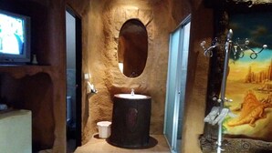 Casa de banho com parede de pedra, espelho redondo, lavatório e pintura representando uma paisagem desértica.