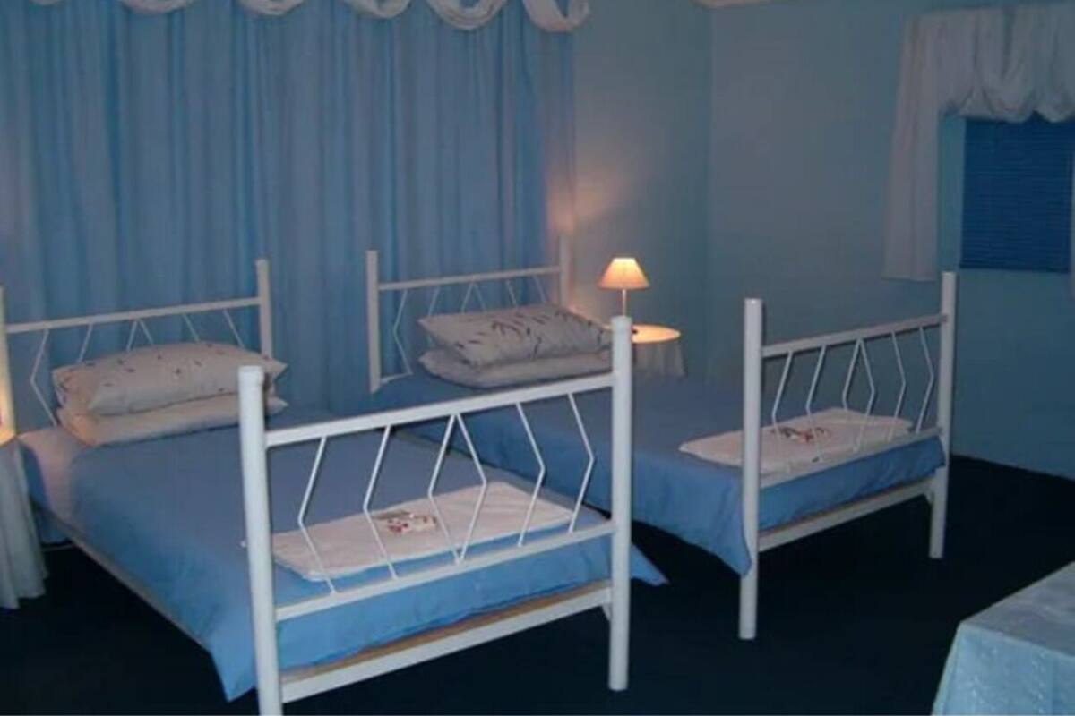 A room with two single beds, each with a white metal frame and blue bedding. There is a nightstand with a lamp on one bed.