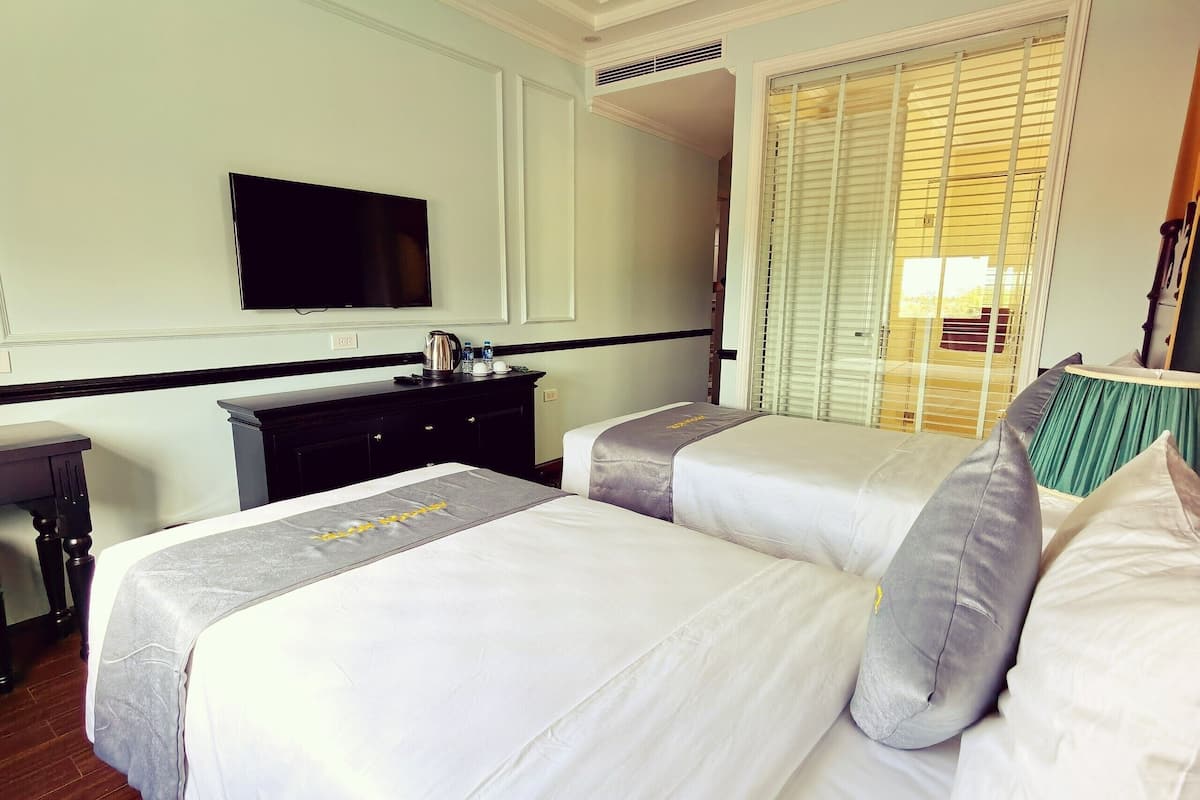 Habitación de hotel con dos camas, un televisor de pantalla plana montado en la pared, una mesita con un hervidor de agua y una ventana con persianas.