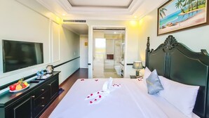 A hotel room with a large bed, a television, a desk with a computer, and a painting of a beach scene. - Dragon Hotel (Bai Chay)
