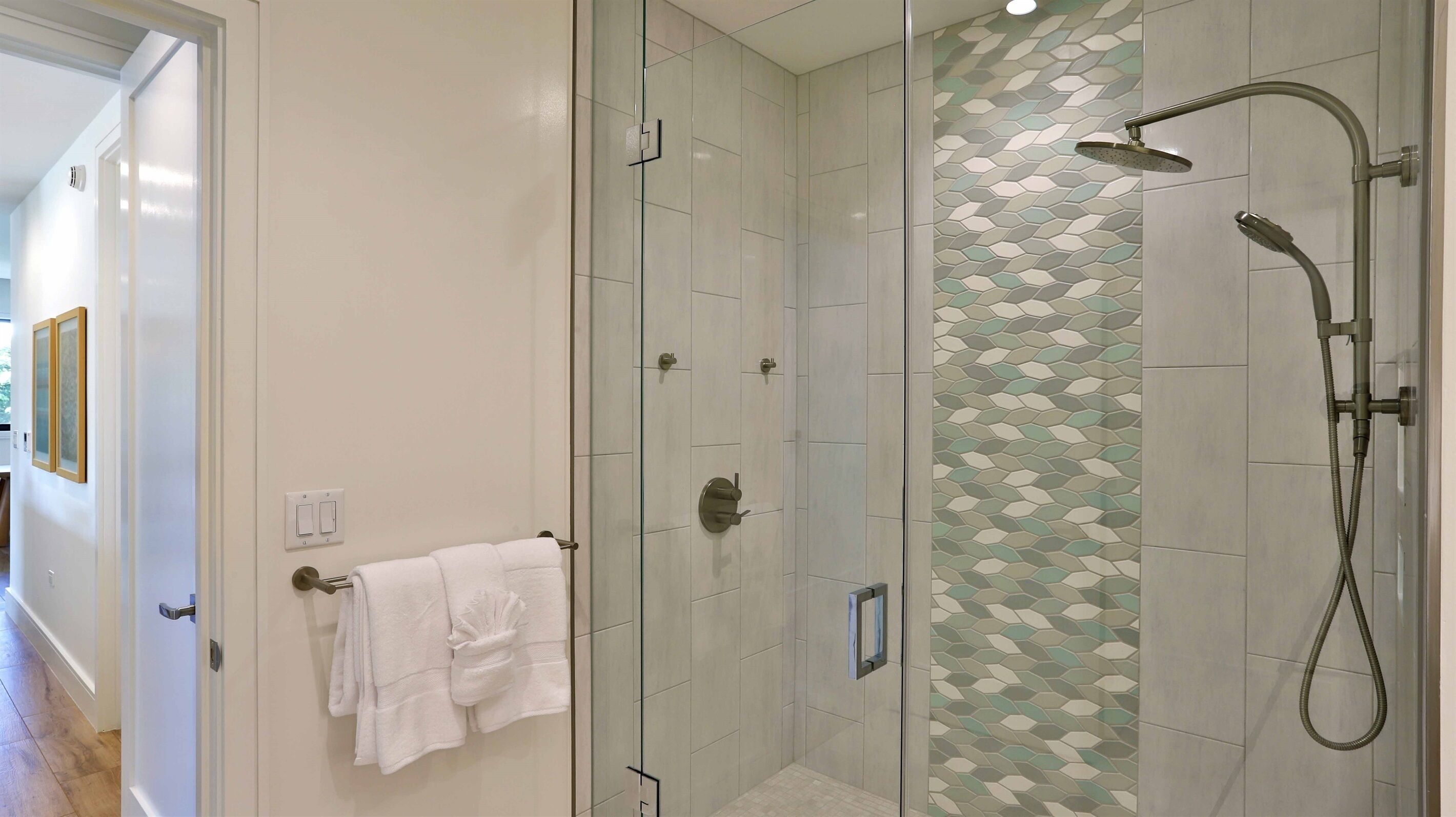 Un baño moderno con ducha de vidrio, pared con diseño de hojas y toallero con toallas blancas.
