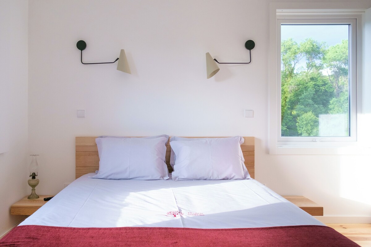 A bedroom with a wooden headboard, a bed with white bedding, two wall-mounted lamps, and a window with a view of trees.