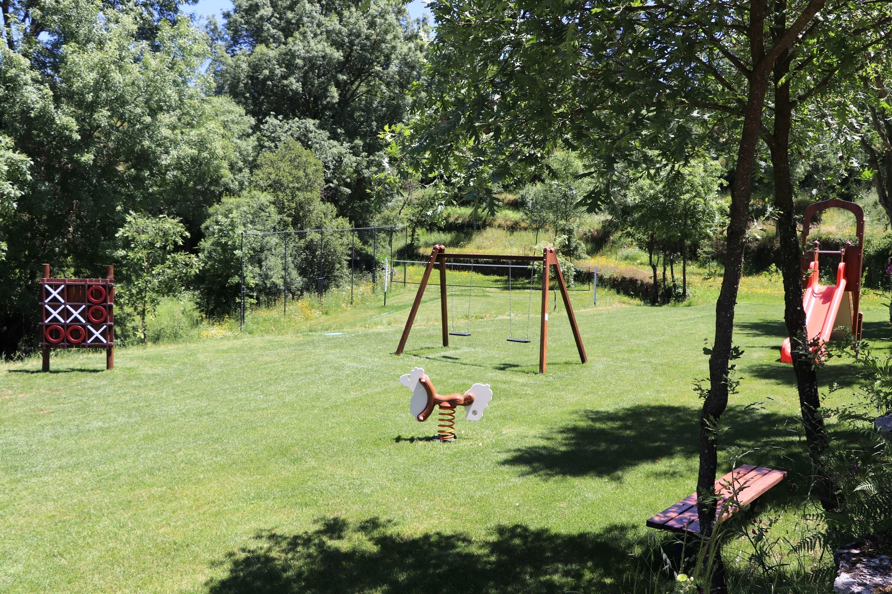 Un parque infantil con columpios, tobogán y estructura para escalar en un área cubierta de césped rodeada de árboles.
