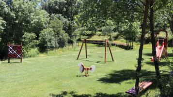 Un parque infantil con columpios, tobogán y estructura para escalar en un área cubierta de césped rodeada de árboles.
