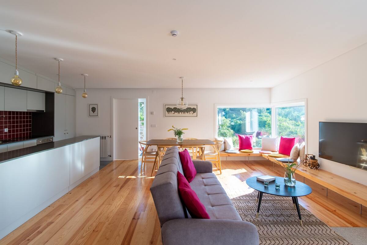 A modern living room with a sofa, coffee table, and a TV. There is a dining area with a table and chairs. The room has wooden flooring and large windows.