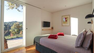 A bedroom with a bed, a TV, a painting, and a view of a forest through a large window.