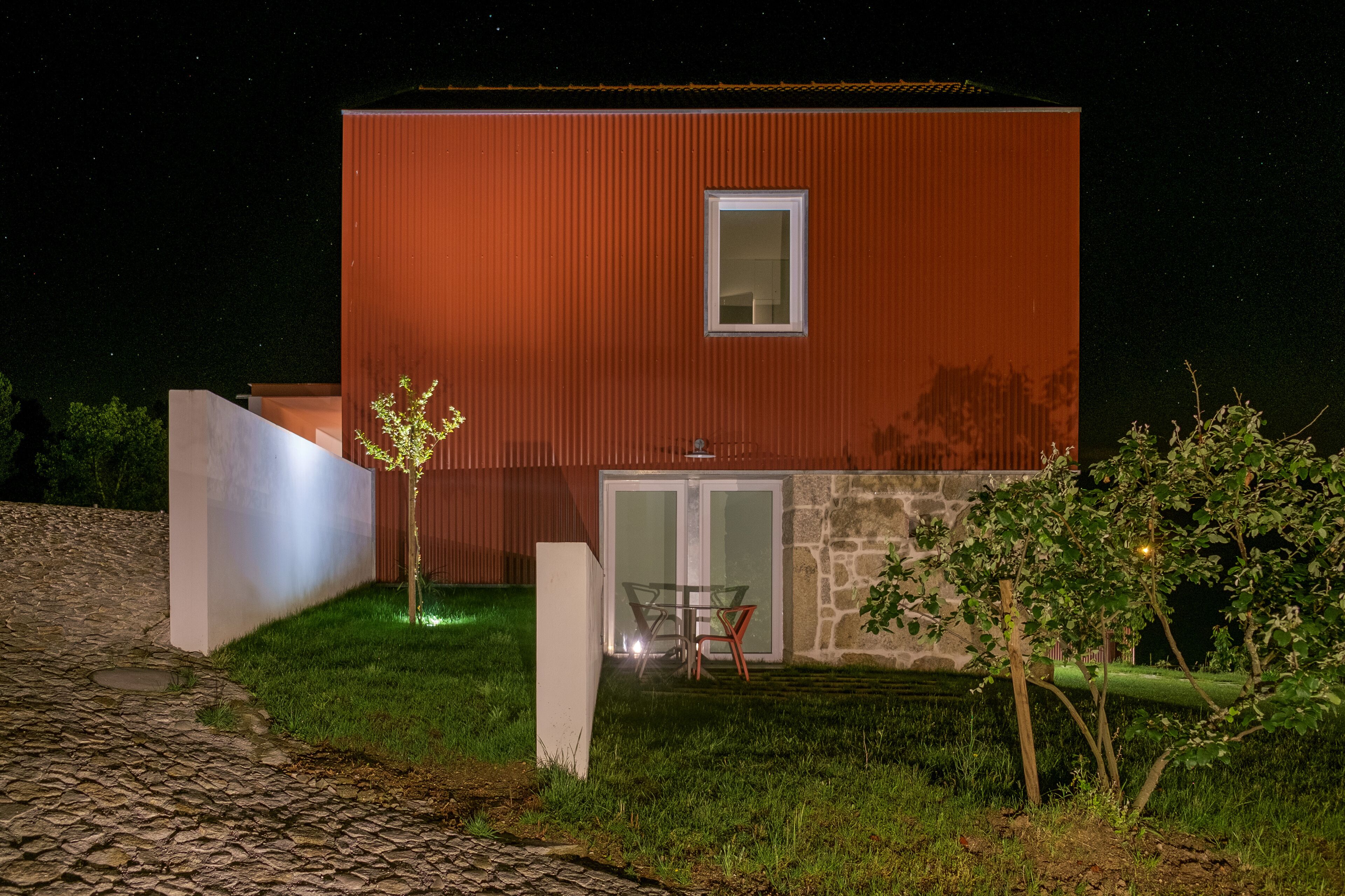 Un edificio moderno con fachada roja, una ventana pequeña y una puerta. Hay un área pavimentada con un árbol y una mesa con sillas.