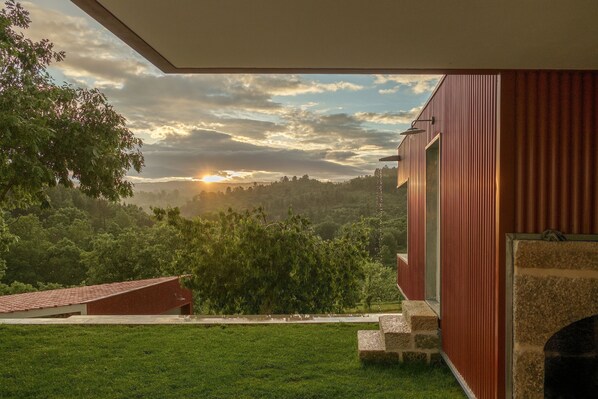 A modern building with a red exterior, a stone fireplace, and a view of a lush green landscape with trees and a sunset. - Encosta do Sobreiro - Serra da Estrela (Fornos de Algodres)