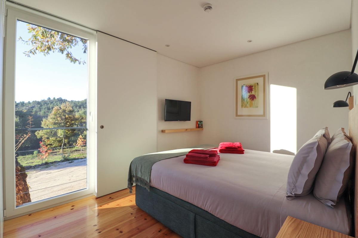 Quarto com cama, televisão, quadro e vista para uma floresta através de uma janela grande.