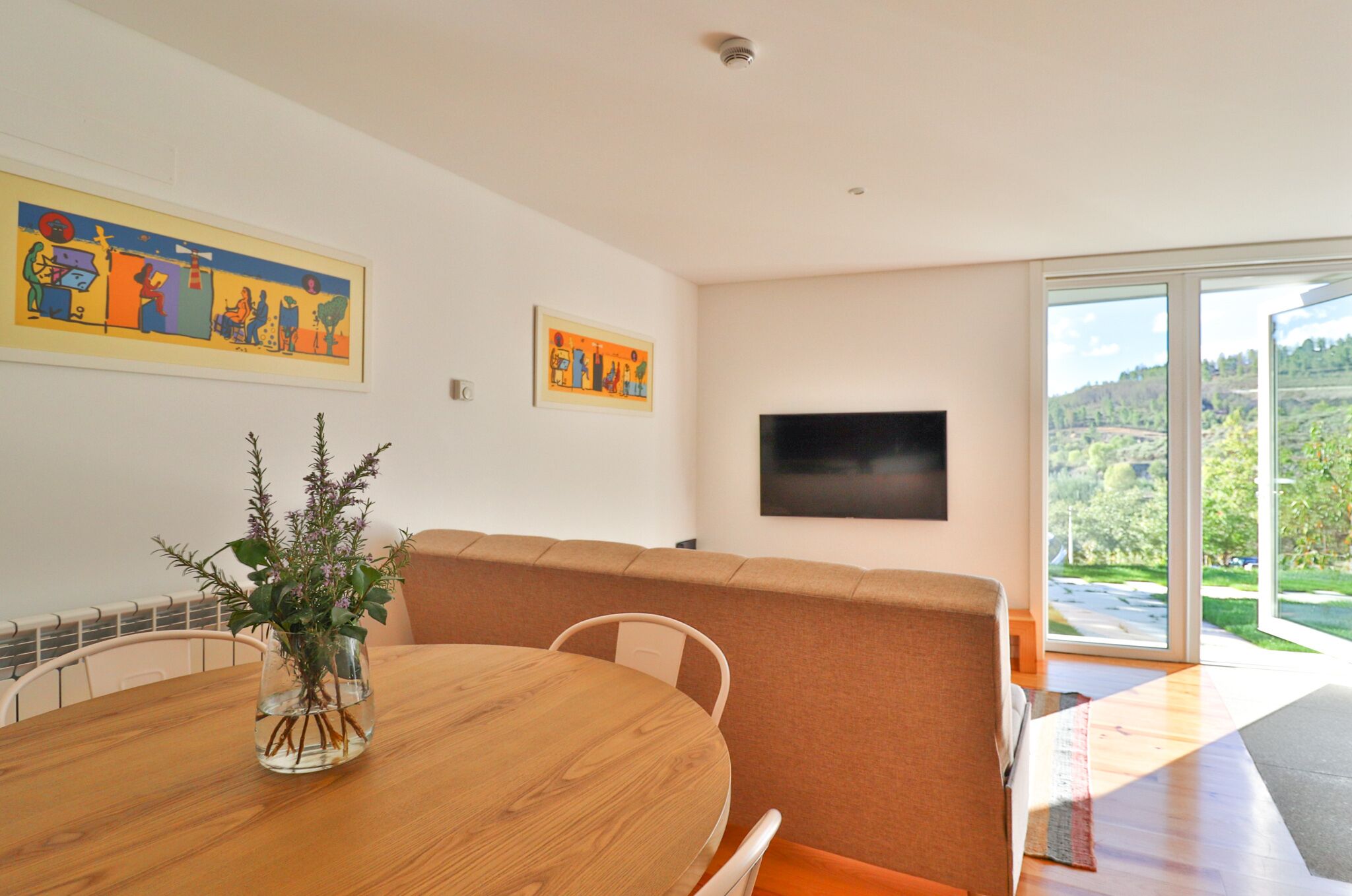 A modern living room with a wooden dining table, a sofa, and a TV mounted on the wall.