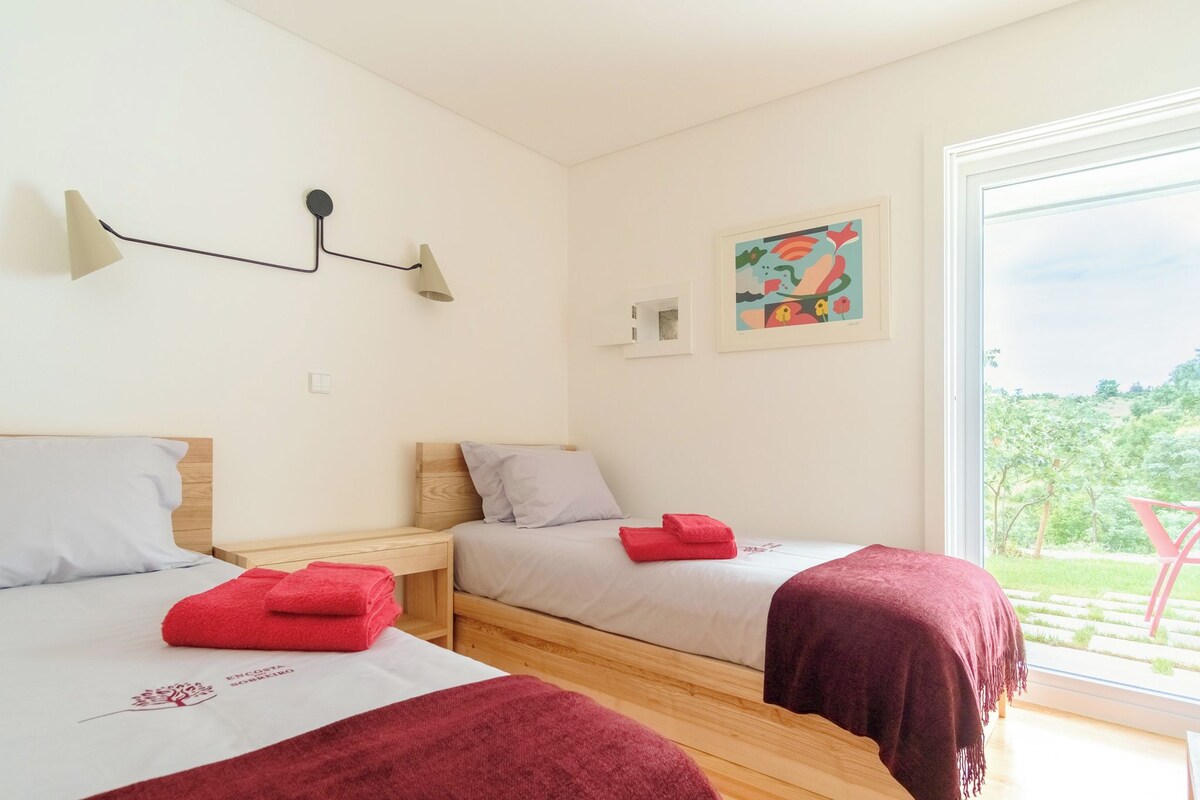 A room with two beds, a window with a view of greenery, a wall-mounted lamp, and a framed artwork.