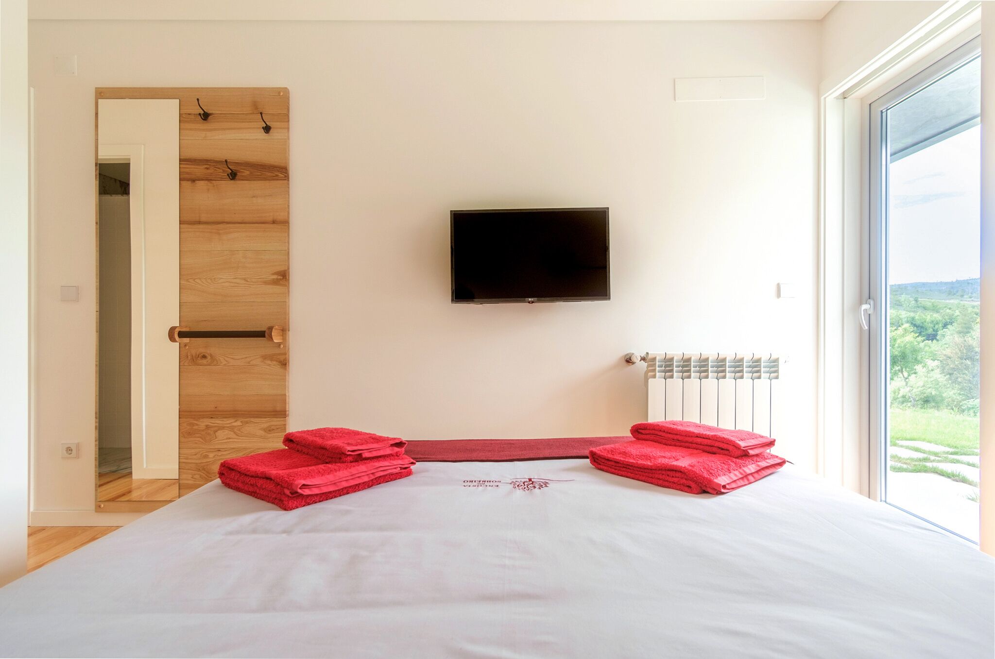 Quarto com cama, televisão na parede, radiador e vista para o verde lá fora.