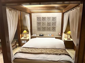 Una habitación de hotel con una cama grande, dos mesitas de noche, una pared con azulejos decorativos y cortinas translúcidas.