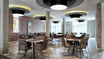 Une salle à manger spacieuse avec de hauts plafonds, des luminaires circulaires et un mur en briques. Des tables et des chaises sont disposées pour les repas.