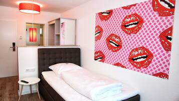 A bedroom with a bed, a bedside table, a mirror, and a wall with a red lips pattern.