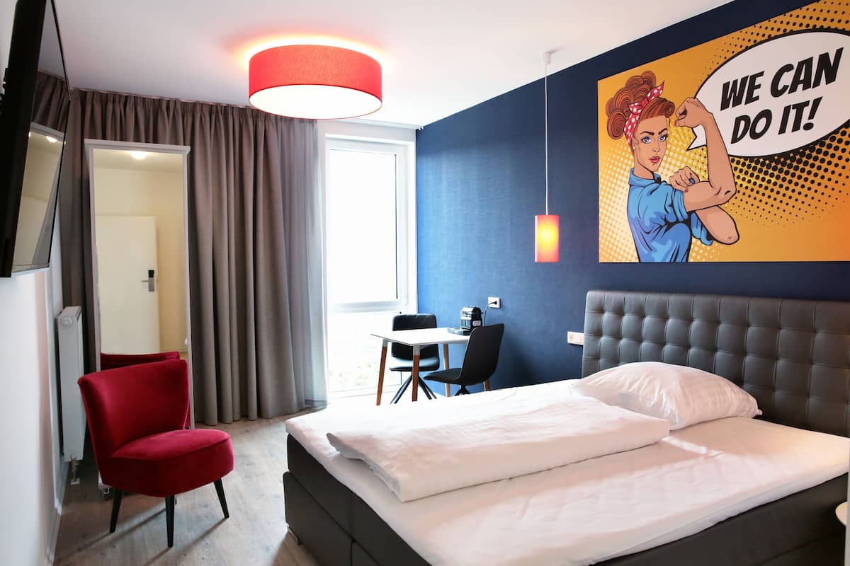Ein Hotelzimmer mit einem Bett, einem roten Sessel, einem Schreibtisch mit Stuhl und einem großen Motivationsplakat an der Wand.