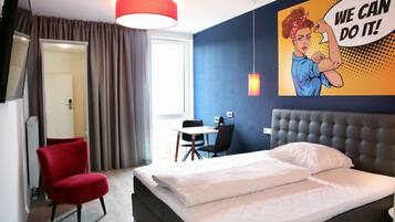 A hotel room with a bed, a red chair, a desk with a chair, and a large motivational poster on the wall.