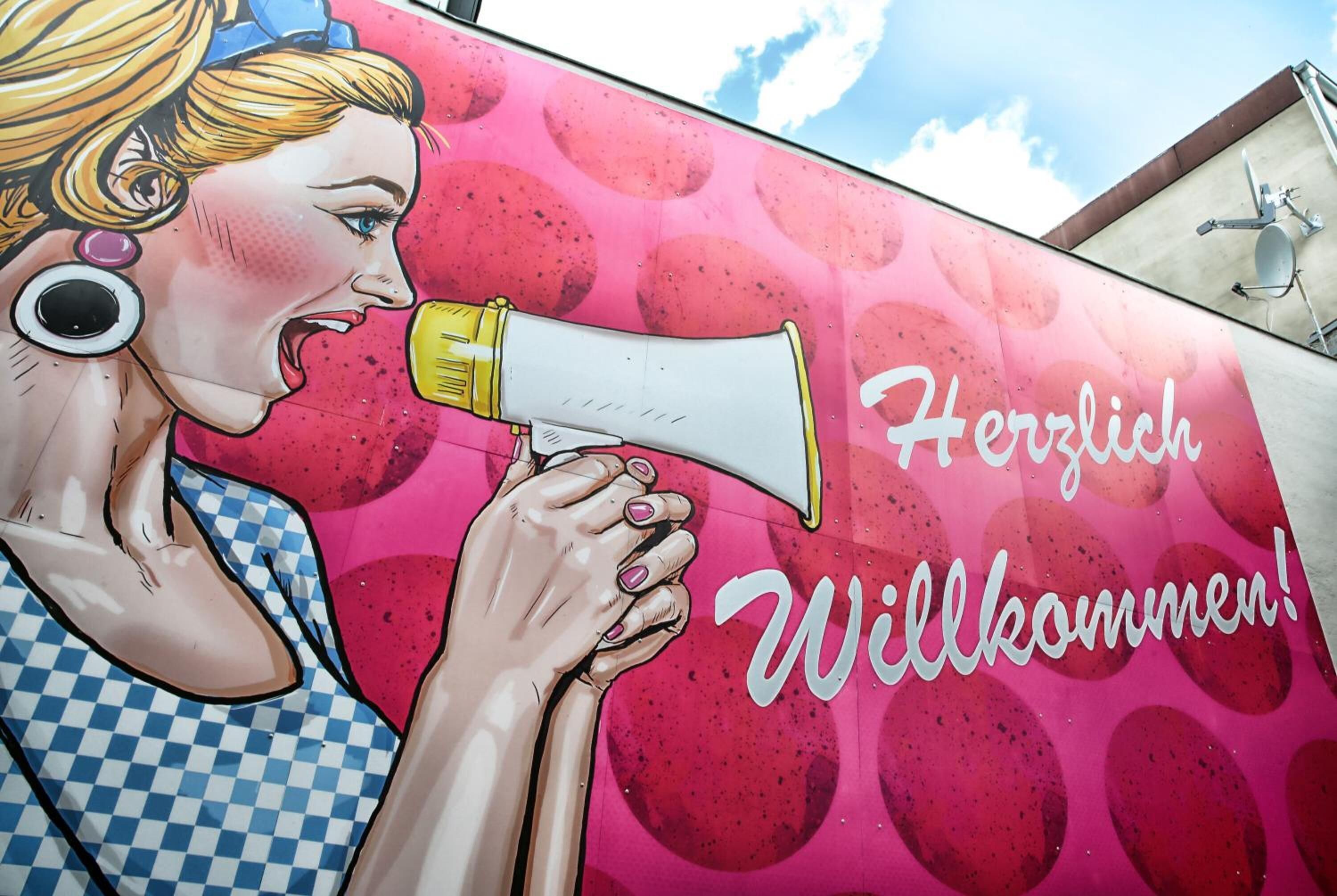 A colorful mural with a woman holding a megaphone and the text "Herzlich Willkommen!"
