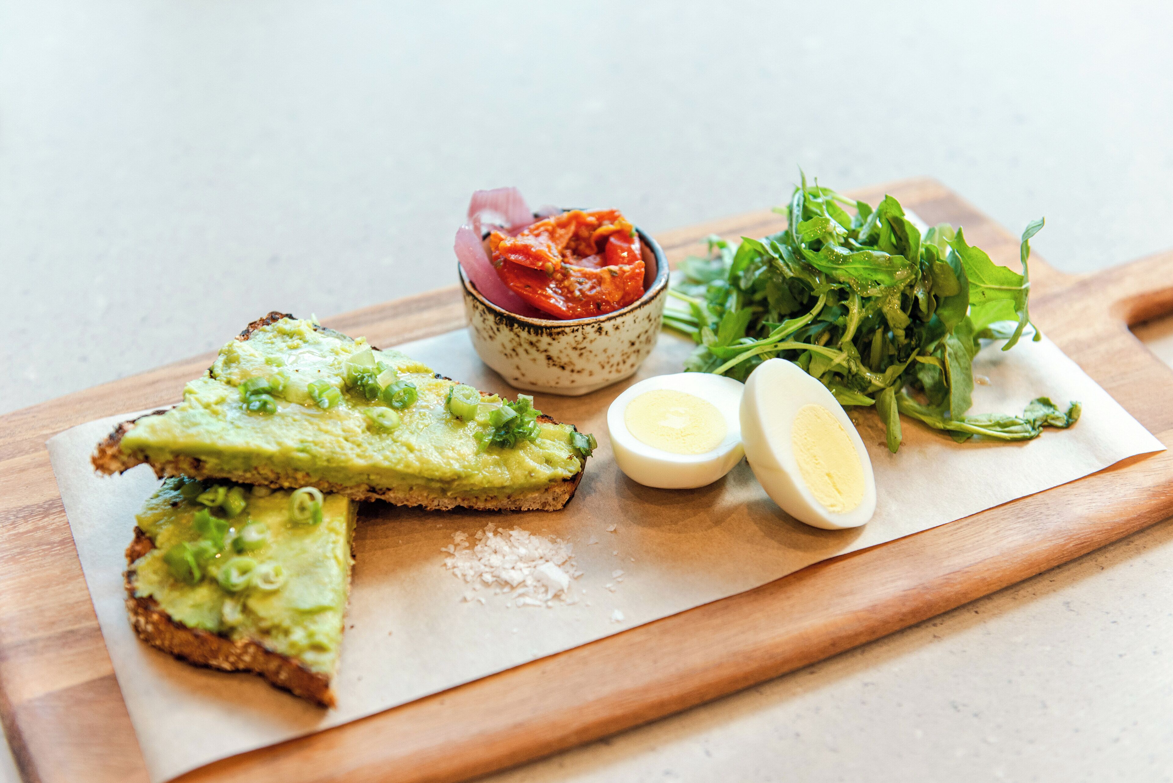 Una tabla de madera con una tostada de aguacate, un bol de salsa roja, un huevo duro y una ensalada de hojas verdes.