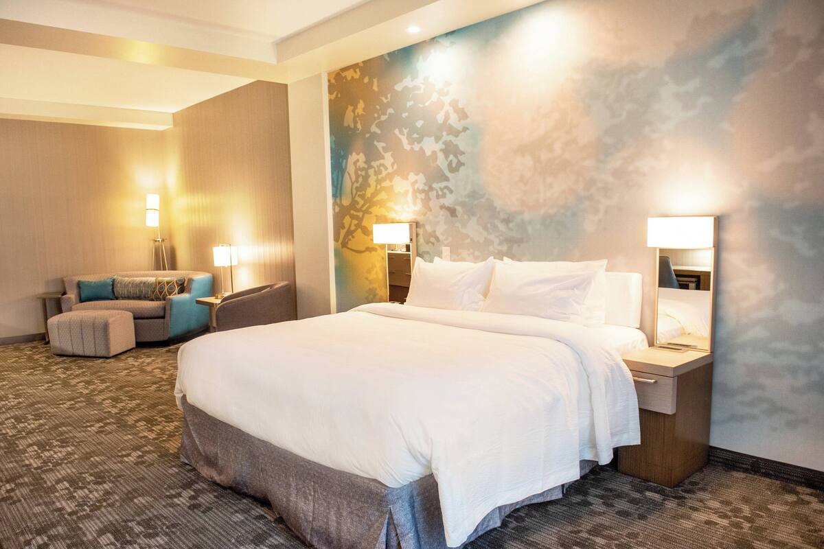 Ein Hotelzimmer mit einem großen Bett, zwei Nachttischen mit Lampen, einer Sitzecke mit Sofa und Hocker sowie einem Wandbild.