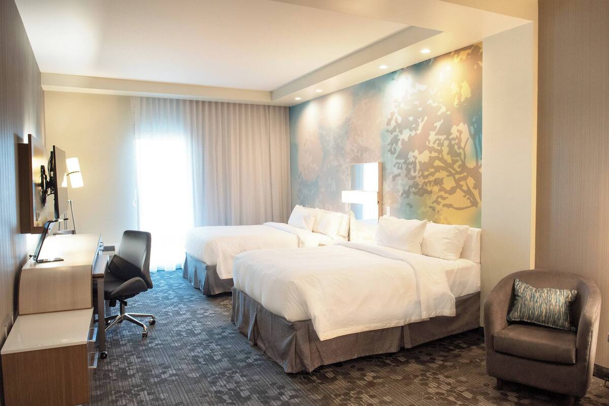 Ein Hotelzimmer mit zwei Betten, einem Schreibtisch, einem Stuhl und einem großen Fenster mit Vorhängen.