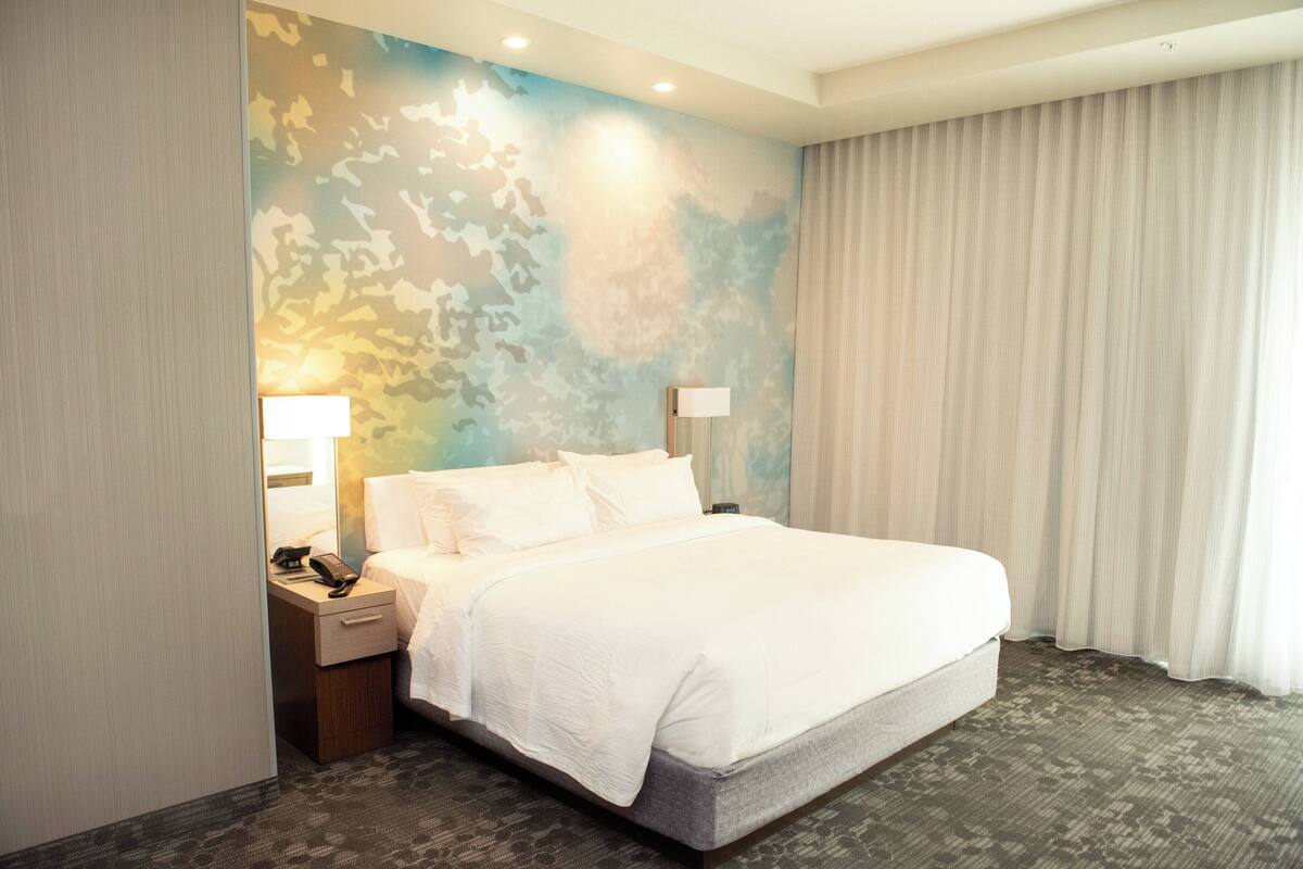 Ein Hotelzimmer mit einem großen Bett, einem Nachttisch mit Lampe, einem Telefon und einer Wand mit Tarnmuster.