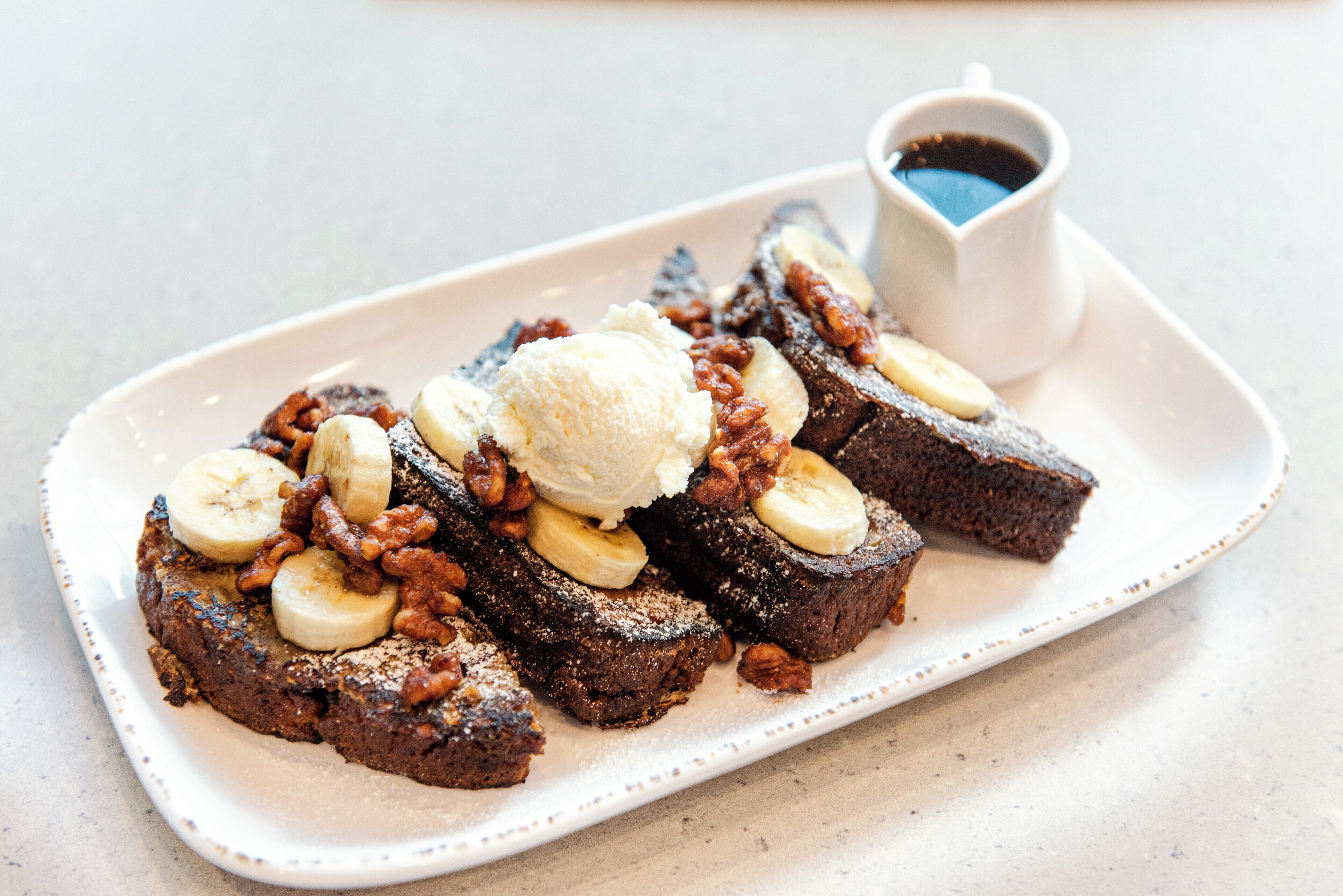 A plate of banana bread with banana slices and a scoop of ice cream on top, accompanied by a small cup of syrup.