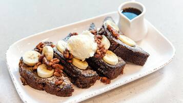 A plate of banana bread with banana slices and a scoop of ice cream on top, accompanied by a small cup of syrup.