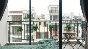 A balcony with a bed, a table, and chairs, overlooking a cityscape with buildings and a clear sky.