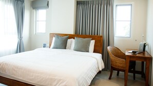 Ein Hotelzimmer mit einem großen Bett, einem hölzernen Kopfteil, einem Korbsessel, einem kleinen Holztisch mit einem Laptop und zwei mit Vorhängen versehenen Fenstern.
