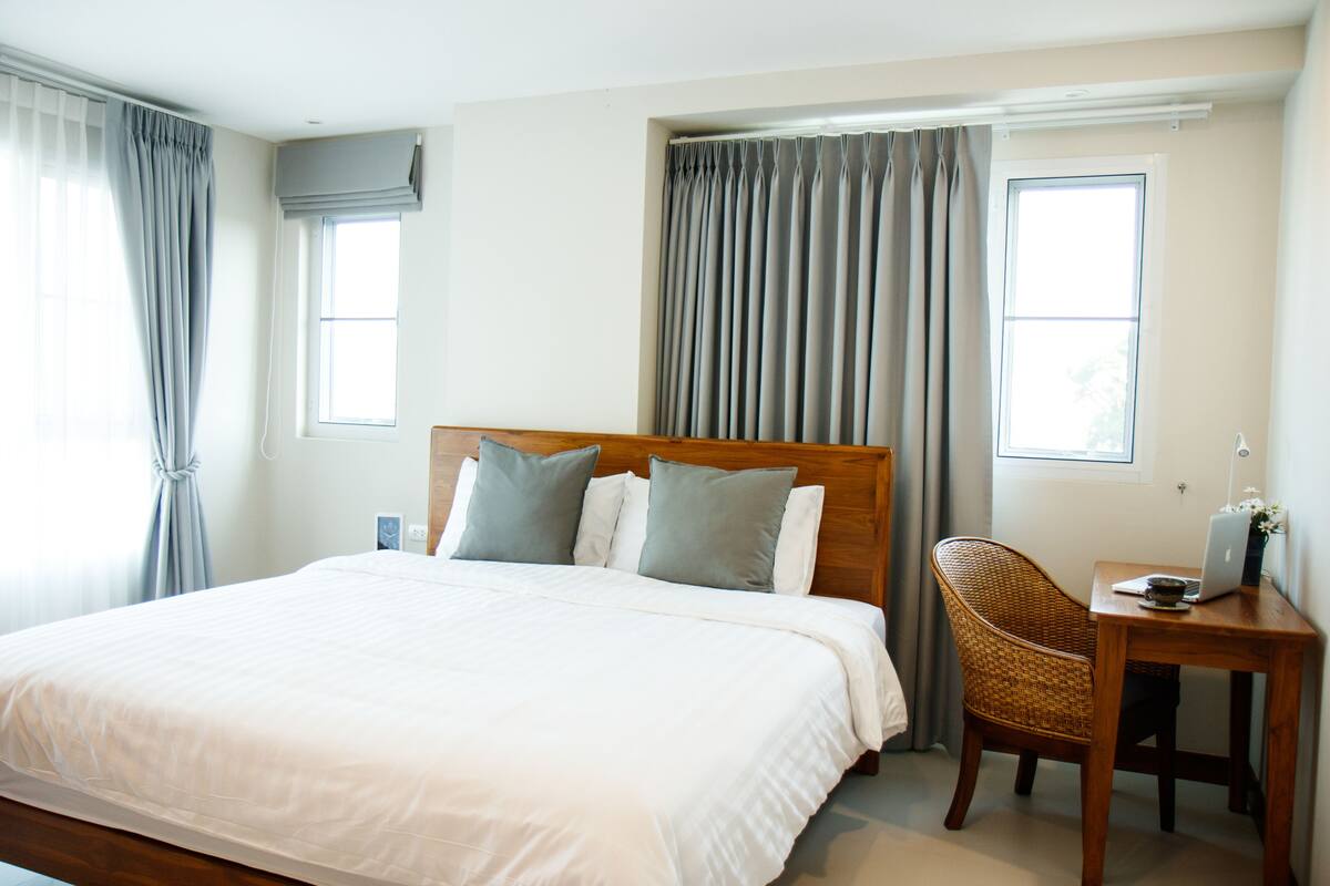 A hotel room with a large bed, a wooden headboard, a wicker chair, a small wooden table with a laptop, and two windows with curtains.