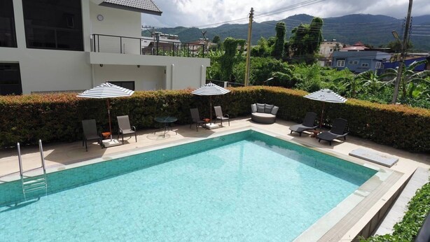 Ein rechteckiger Pool mit Liegestühlen und Sonnenschirmen, umgeben von einer Hecke und einem Gebäude mit Balkon.