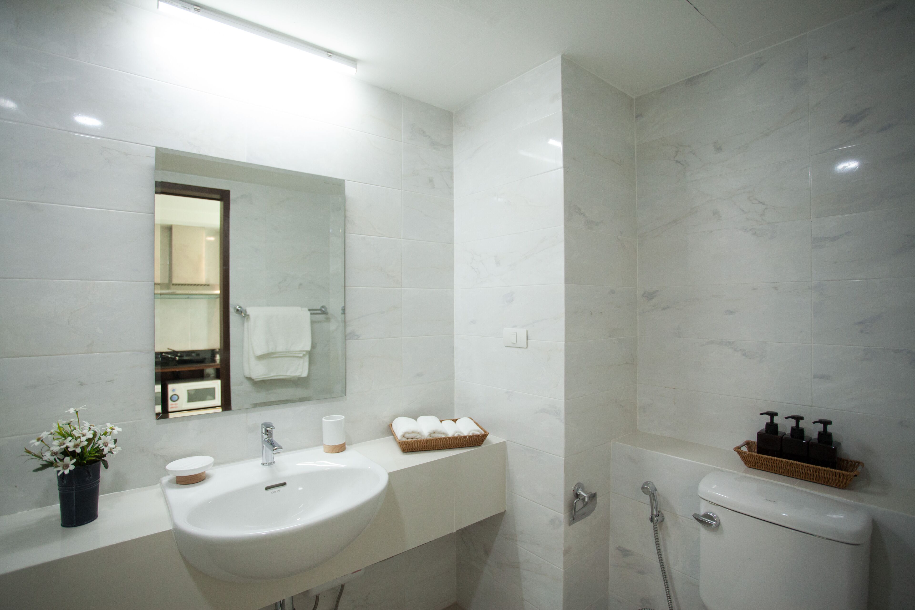 Ein modernes Badezimmer mit einem weißen Waschbecken, Spiegel und Toilette. Auf der Ablage liegen Pflegeprodukte und ein Korb mit Handtüchern.