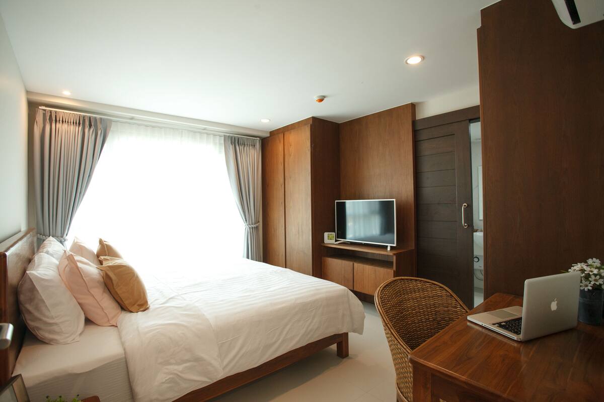 A hotel room with a large bed, a television on a wooden stand, a laptop on a wooden table, and a wicker chair.